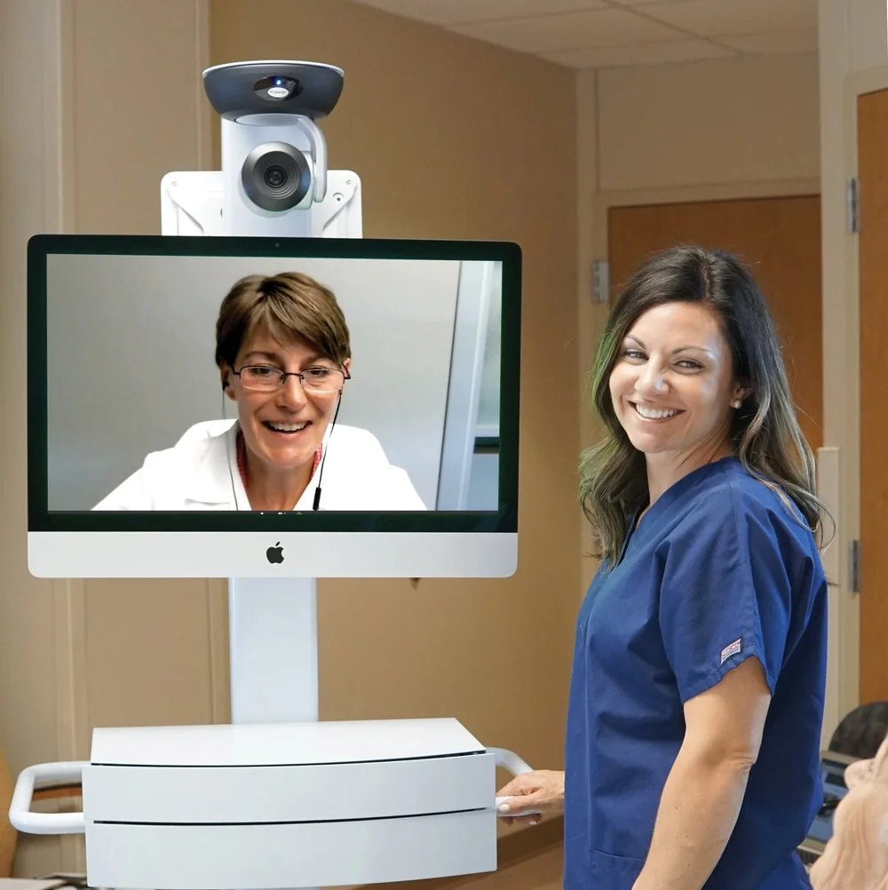 A higher-grade doctor consulting with a nurse on an Apple monitor screen equipped with a large webcam.